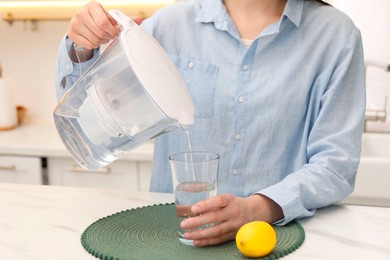 Woman pouring water from filter jug into glass at white marble table in kitchen, closeup