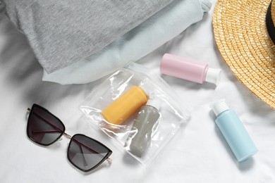 Photo of Cosmetic travel kit. Plastic bag with small containers of personal care products, beach accessories and clothes on bed, above view