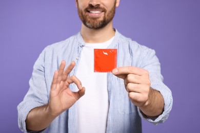 Photo of Man with condom showing ok gesture on purple background, closeup. Safe sex