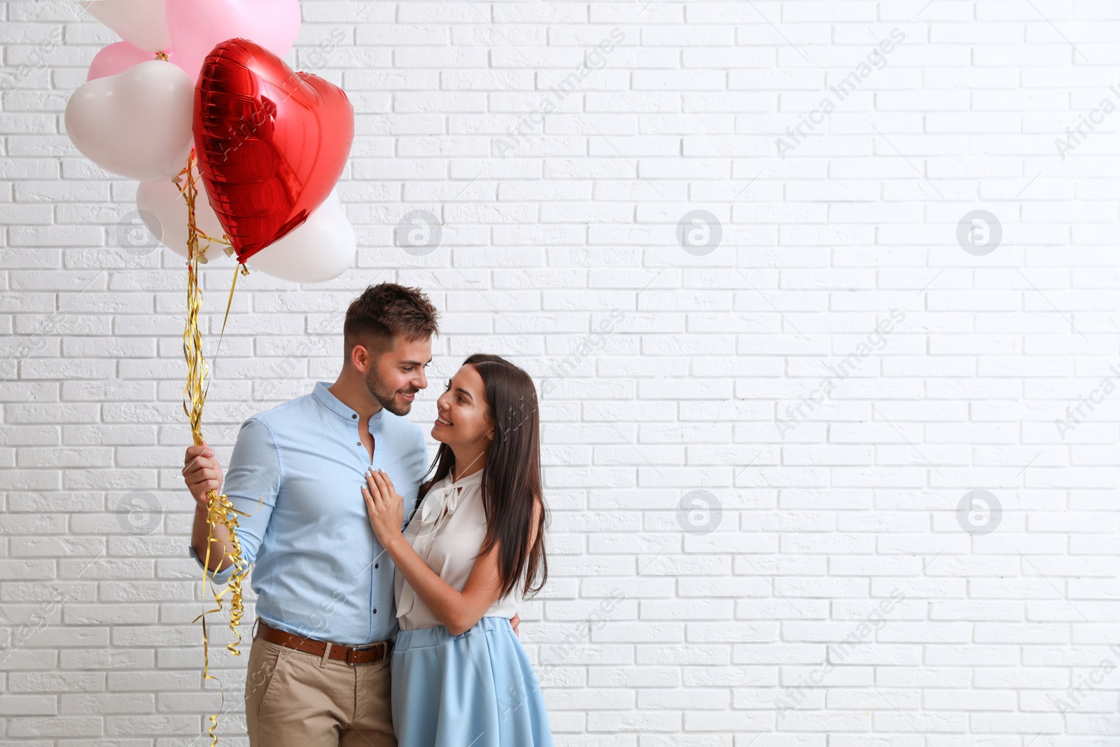 Photo of Young couple with air balloons near white brick wall. Celebration of Saint Valentine's Day