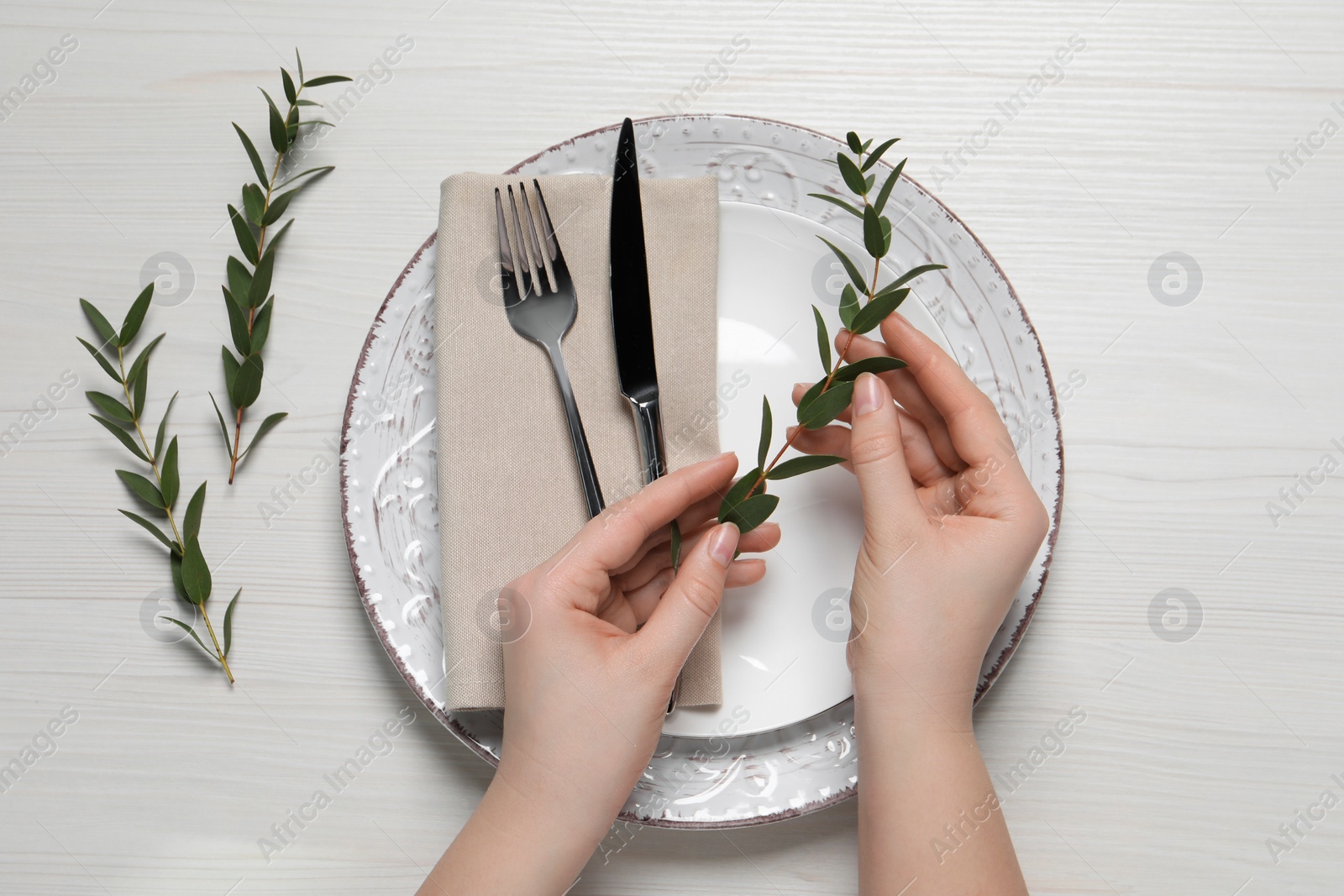 Photo of Woman adjusting stylish table setting with eucalyptus leaves on white wooden background, closeup