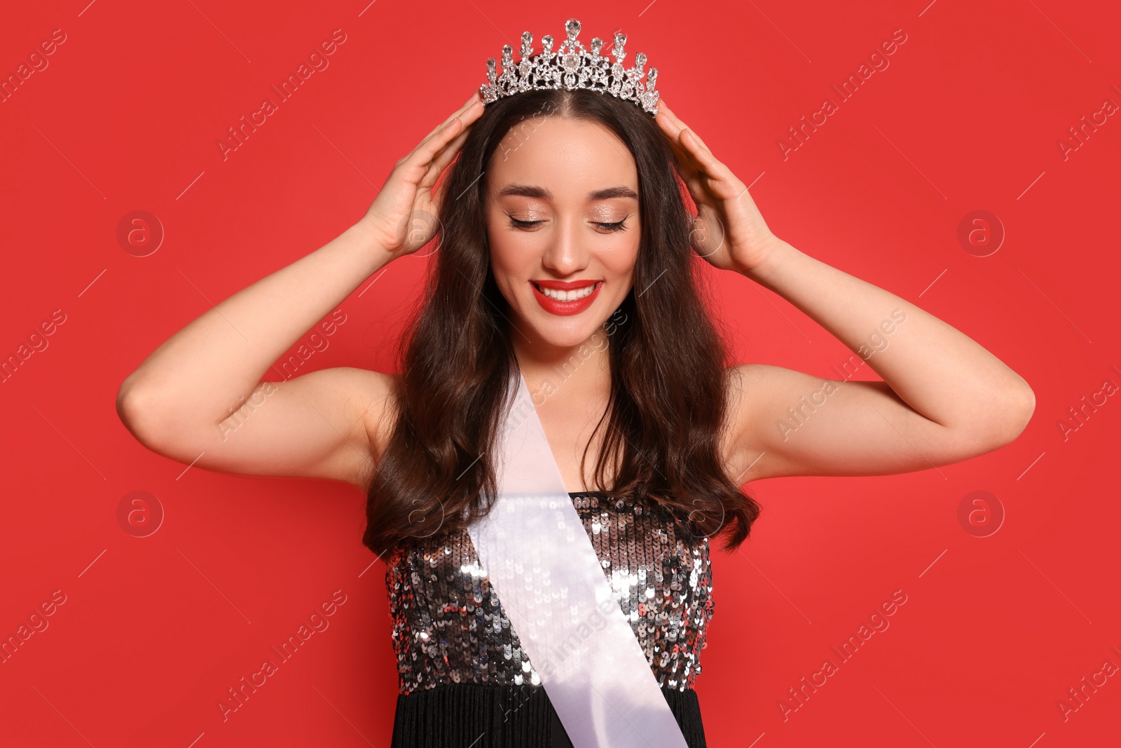 Photo of Beautiful young woman with tiara and ribbon in dress on red background. Beauty contest
