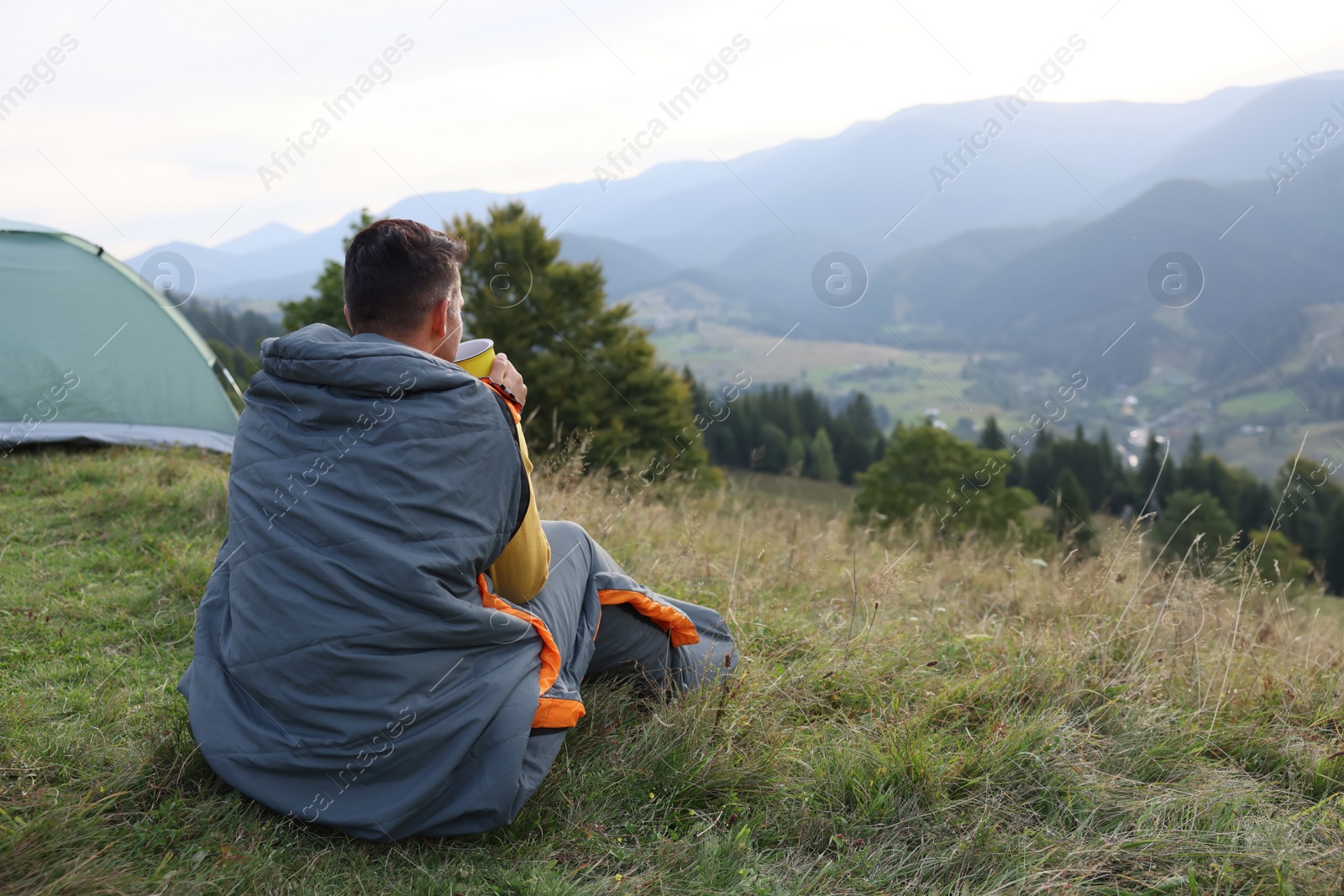 Photo of Man with cup of drink in sleeping bag enjoying mountain landscape, back view. Space for text