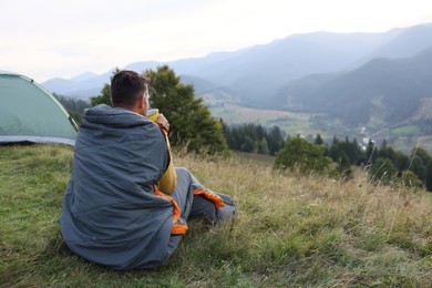 Man with cup of drink in sleeping bag enjoying mountain landscape, back view. Space for text