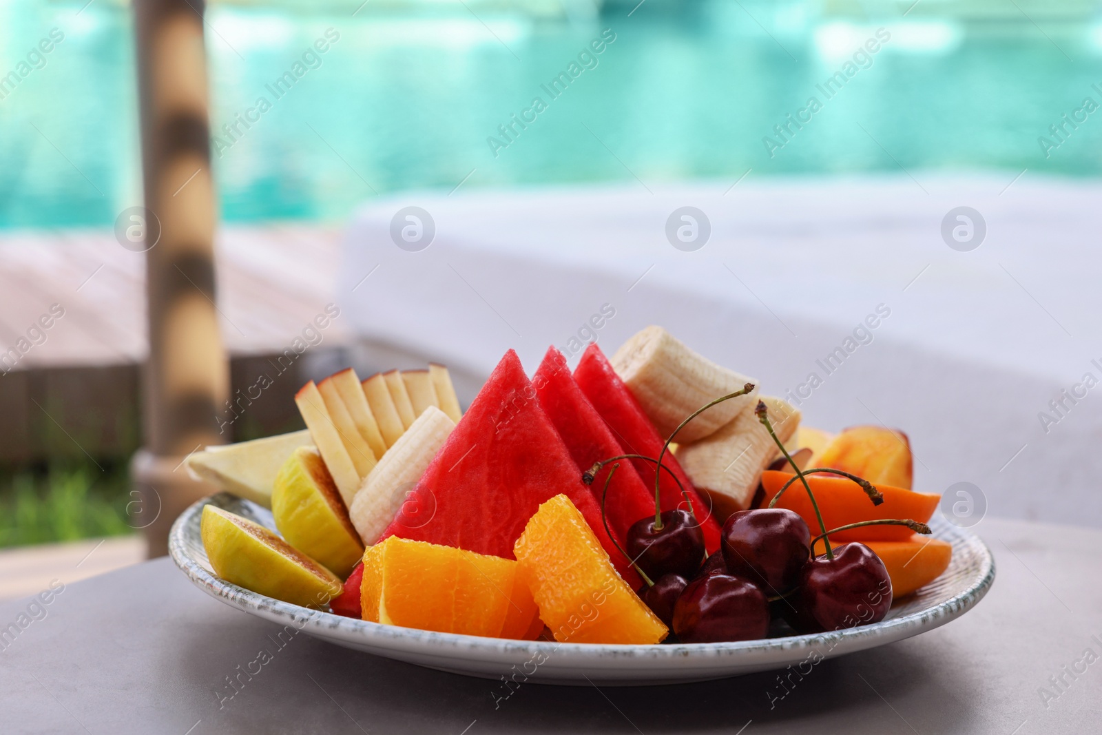 Photo of Plate with fresh fruits on table near sun lounger. Luxury resort with outdoor swimming pool