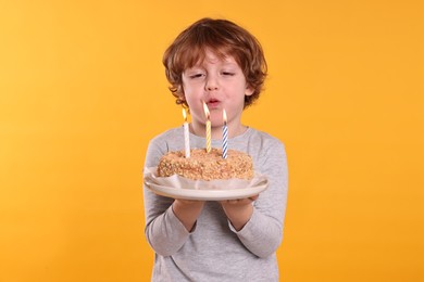 Birthday celebration. Cute little boy blowing candles on tasty cake against orange background