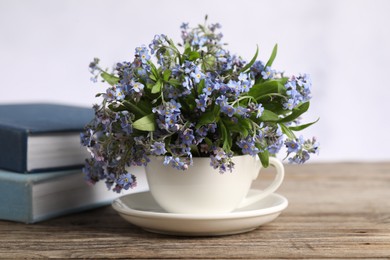 Photo of Beautiful forget-me-not flowers in cup, saucer and books on wooden table against light background, closeup