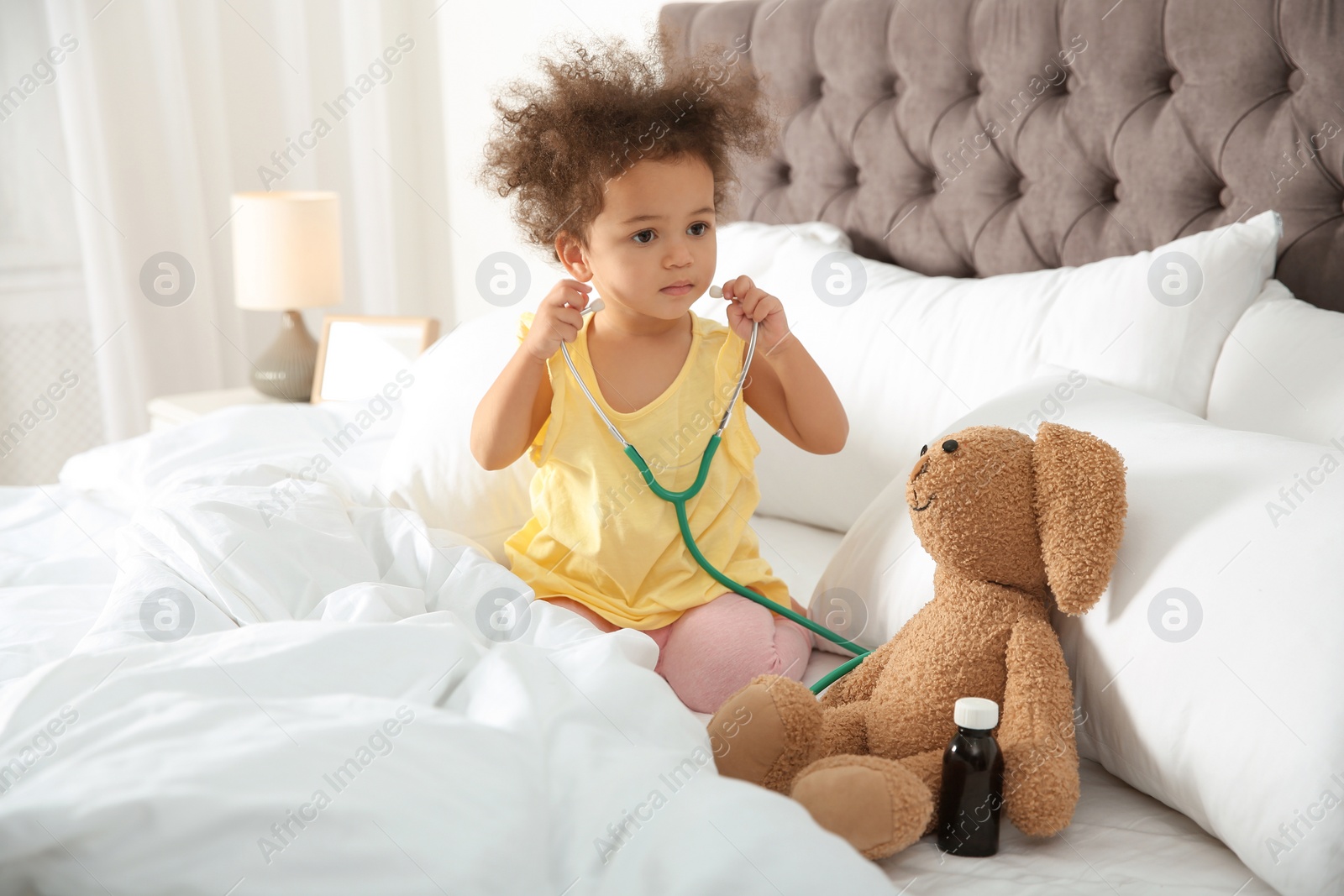 Photo of Cute African American child imagining herself as doctor while playing with stethoscope and toy bunny at home