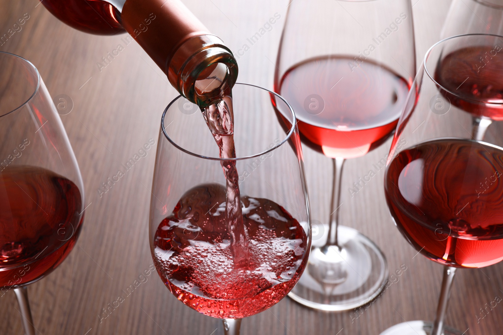 Photo of Pouring rose wine from bottle into glasses on wooden table, closeup