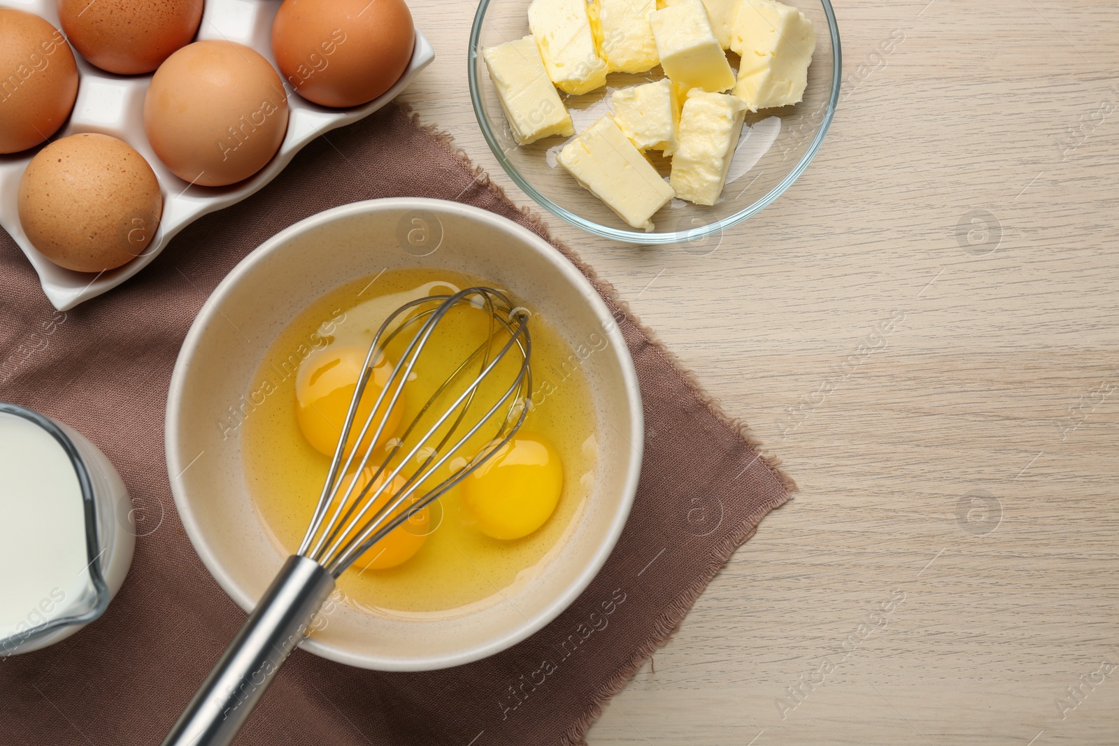 Photo of Whisk and eggs in bowl, butter, milk on wooden table, flat lay. Space for text