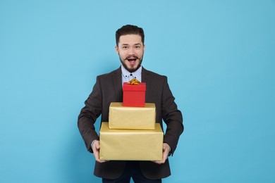Emotional man in suit with stack of gifts on light blue background