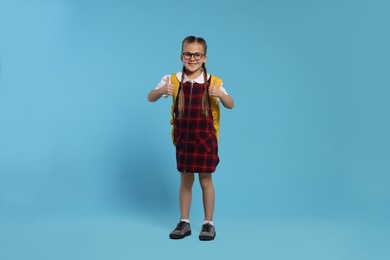 Happy schoolgirl in glasses with backpack showing thumb up gesture on light blue background