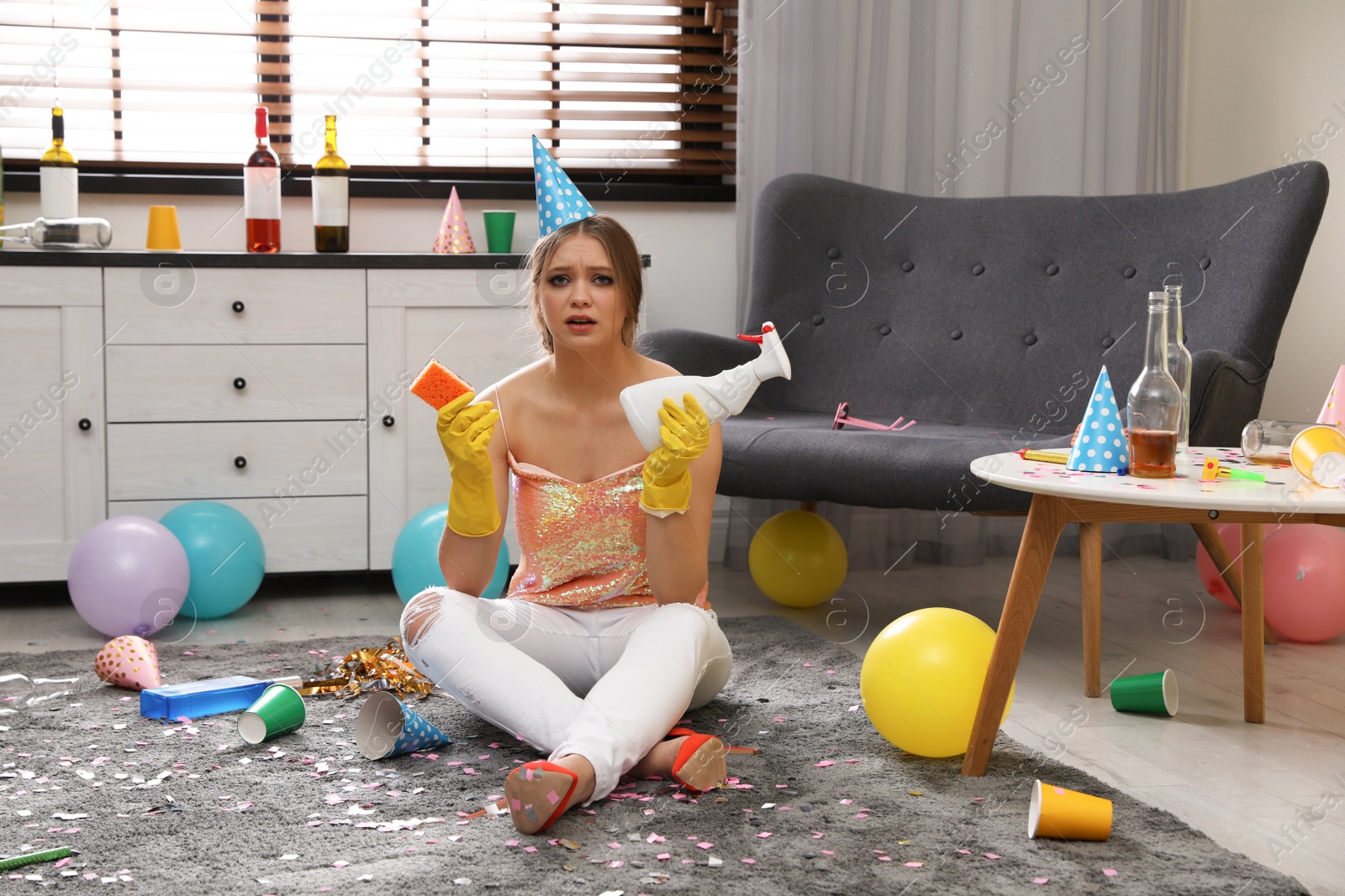 Photo of Tired young woman with bottle of detergent and sponge in messy room after party