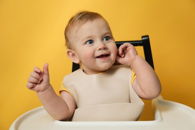 Photo of Cute little baby wearing bib in highchair on yellow background