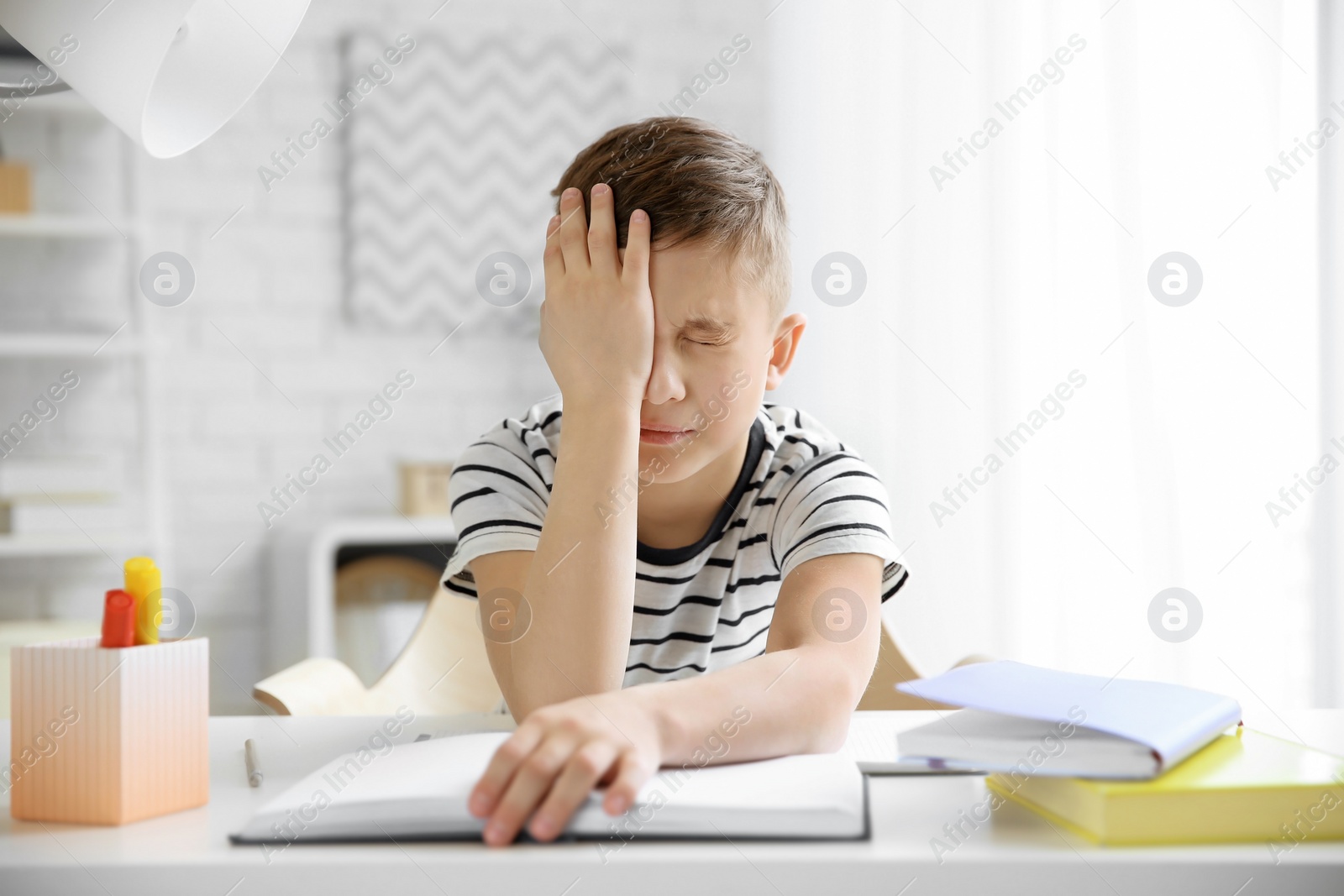 Photo of Little boy suffering from headache while doing homework at table indoors