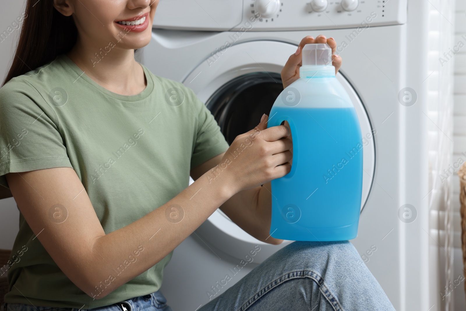 Photo of Woman sitting near washing machine and holding fabric softener, closeup