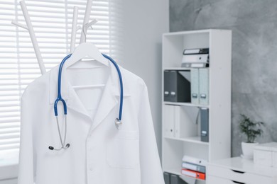 Photo of White doctor's gown and stethoscope hanging on rack in clinic, closeup. Space for text