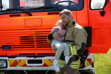Photo of Firefighter in uniform holding rescued little girl near fire truck outdoors. Save life