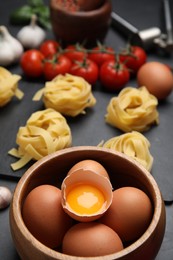 Photo of Raw eggs, uncooked tagliatelle and fresh ingredients on black table, closeup