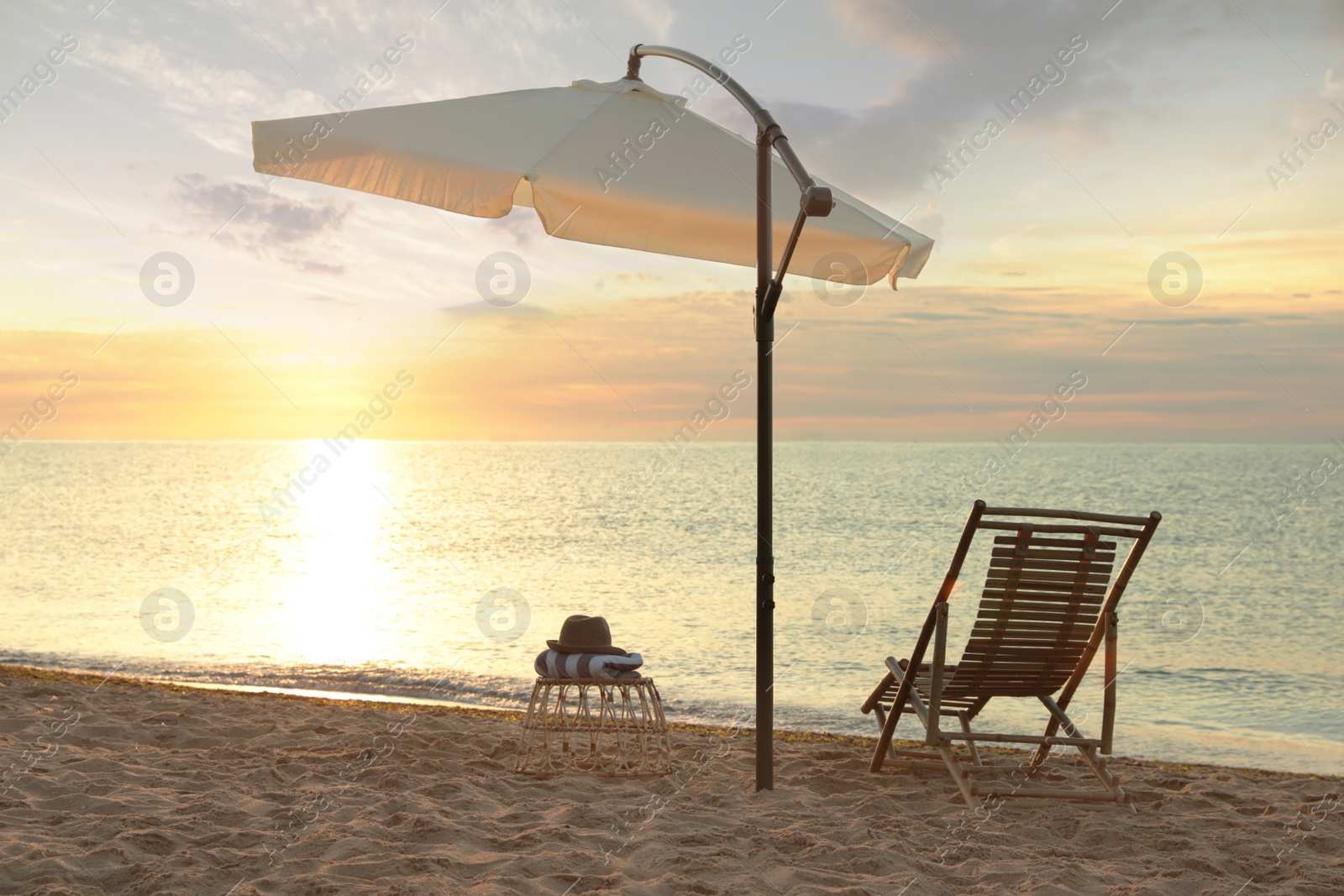 Photo of Wooden deck chair, outdoor umbrella and wicker table on sandy beach at sunset. Summer vacation