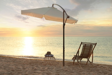 Photo of Wooden deck chair, outdoor umbrella and wicker table on sandy beach at sunset. Summer vacation