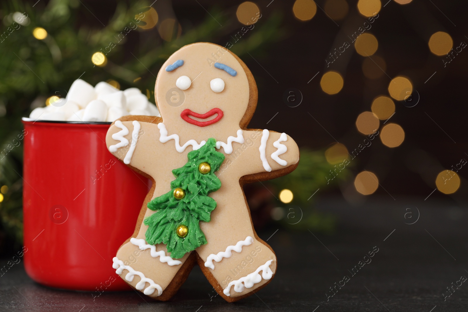 Photo of Gingerbread man and red cup with marshmallows on black table against blurred festive lights. Making homemade Christmas cookies