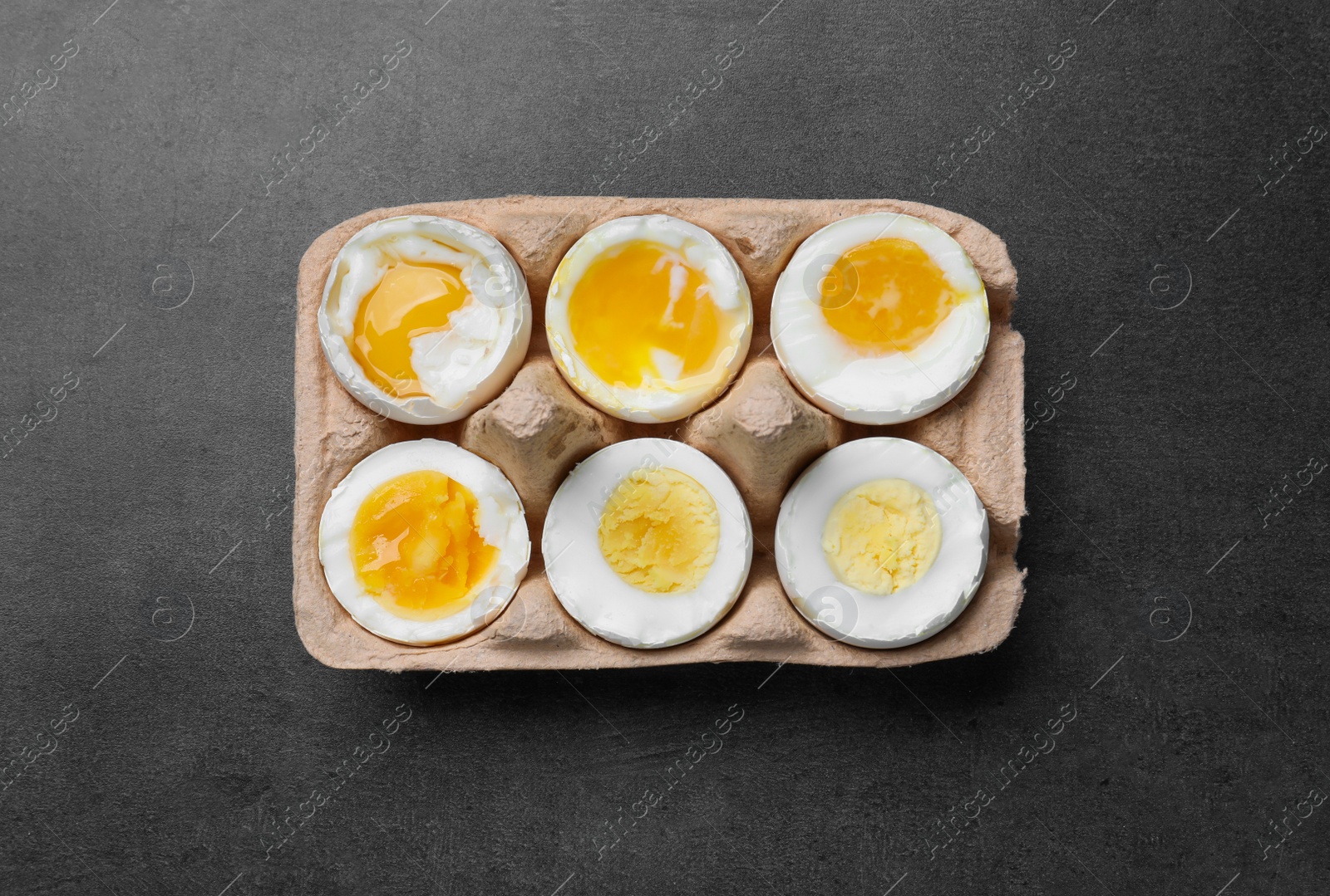 Photo of Boiled chicken eggs of different readiness stages in carton on dark grey table, top view