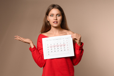 Photo of Emotional young woman holding calendar with marked menstrual cycle days on beige background