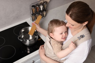 Photo of Mother holding her child in sling (baby carrier) while cooking pasta in pot indoors, above view