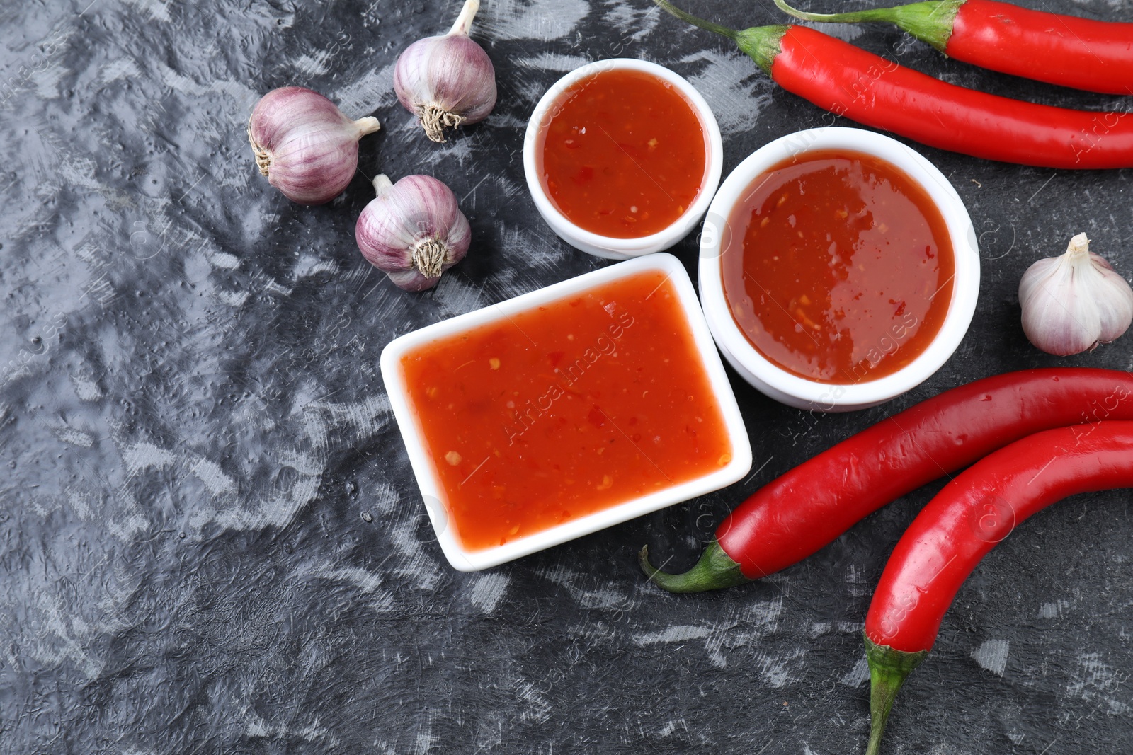 Photo of Spicy chili sauce, peppers and garlic on black textured table, flat lay. Space for text