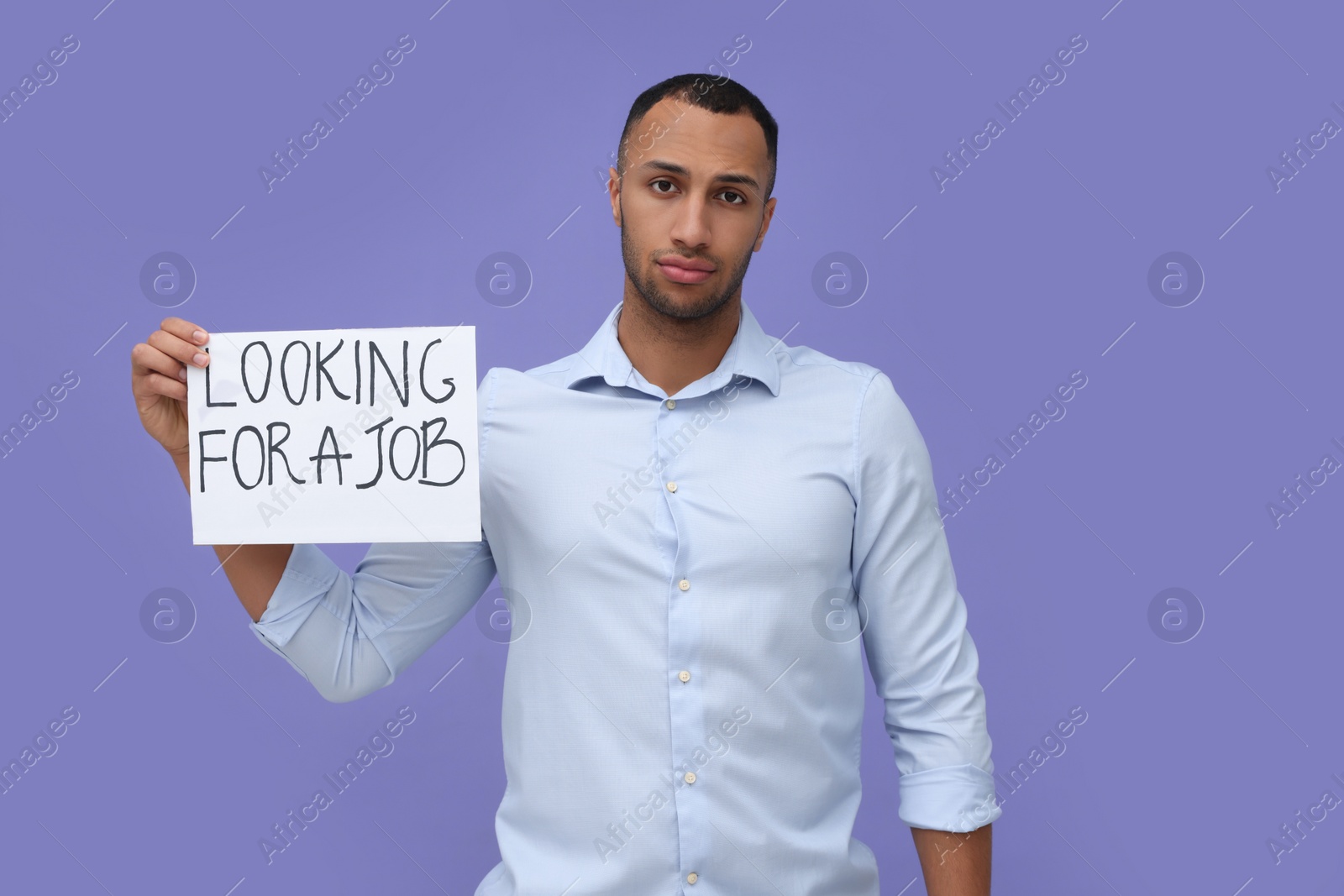 Photo of Young unemployed man holding sign with phrase Looking For A Job on violet background