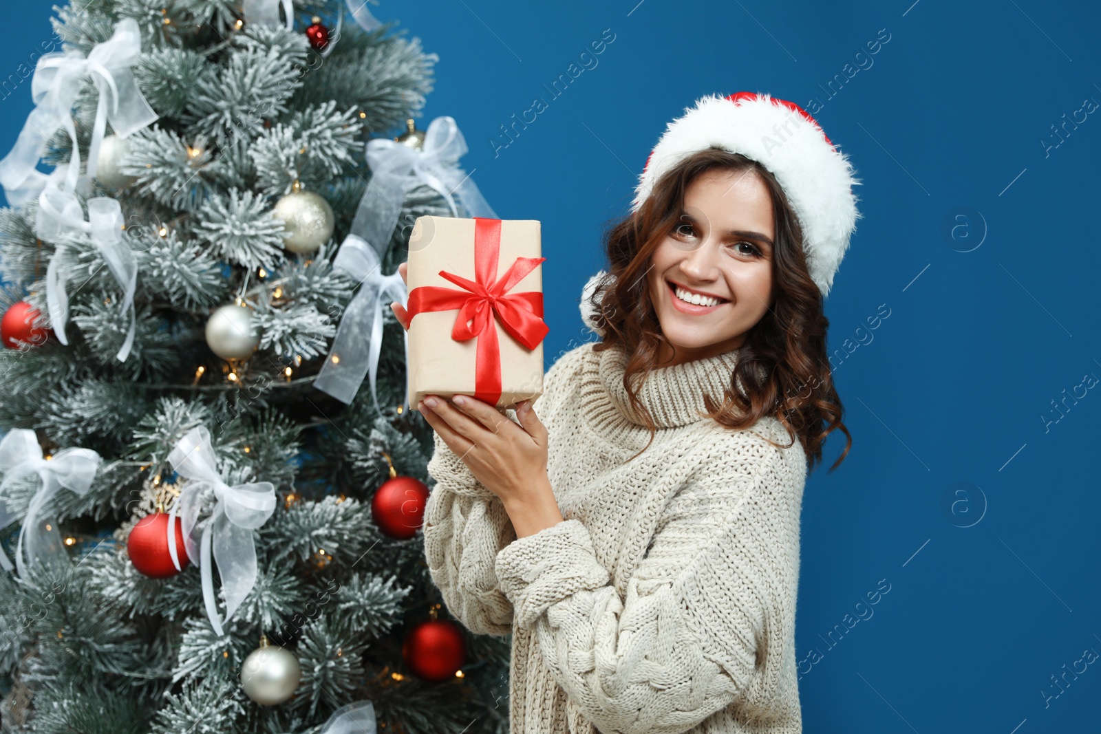 Image of Happy young woman in Santa hat with gift near Christmas tree on blue background