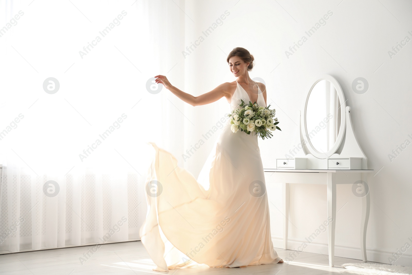 Photo of Young bride in wedding dress with beautiful bouquet near mirror indoors