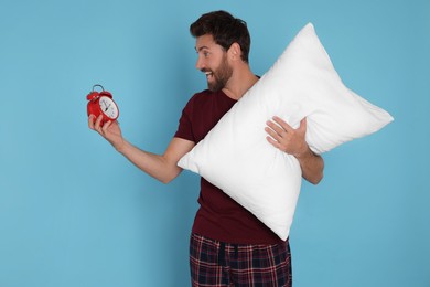 Emotional overslept man with alarm clock and pillow on light blue background