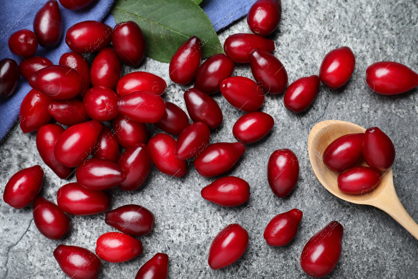 Photo of Pile of fresh ripe dogwood berries with green leaf and wooden spoon on grey table, flat lay