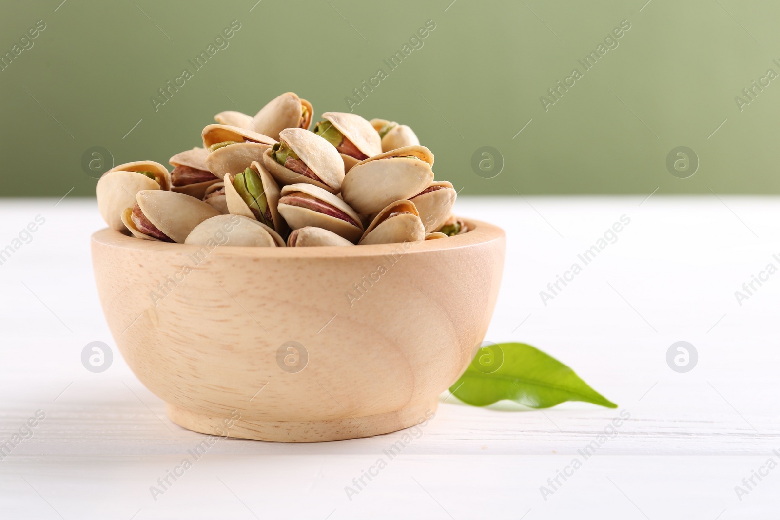 Photo of Tasty pistachios in bowl on white wooden table against olive background, closeup. Space for text