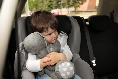 Sad little boy with toy sitting in safety seat alone inside car. Child in danger