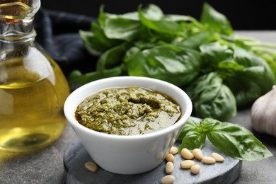 Photo of Tasty pesto sauce in bowl, basil, pine nuts and oil on grey table