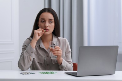 Depressed woman with glass of water taking antidepressant pill at white table indoors