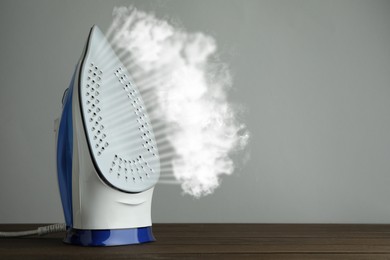 Modern iron with steam on wooden table against light grey background, space for text