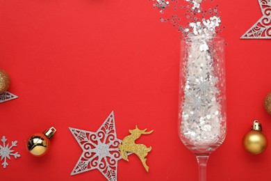 Photo of Flat lay composition with shiny confetti spilled out of champagne glass near Christmas decorations on red background, space for text