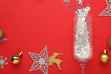 Flat lay composition with shiny confetti spilled out of champagne glass near Christmas decorations on red background, space for text