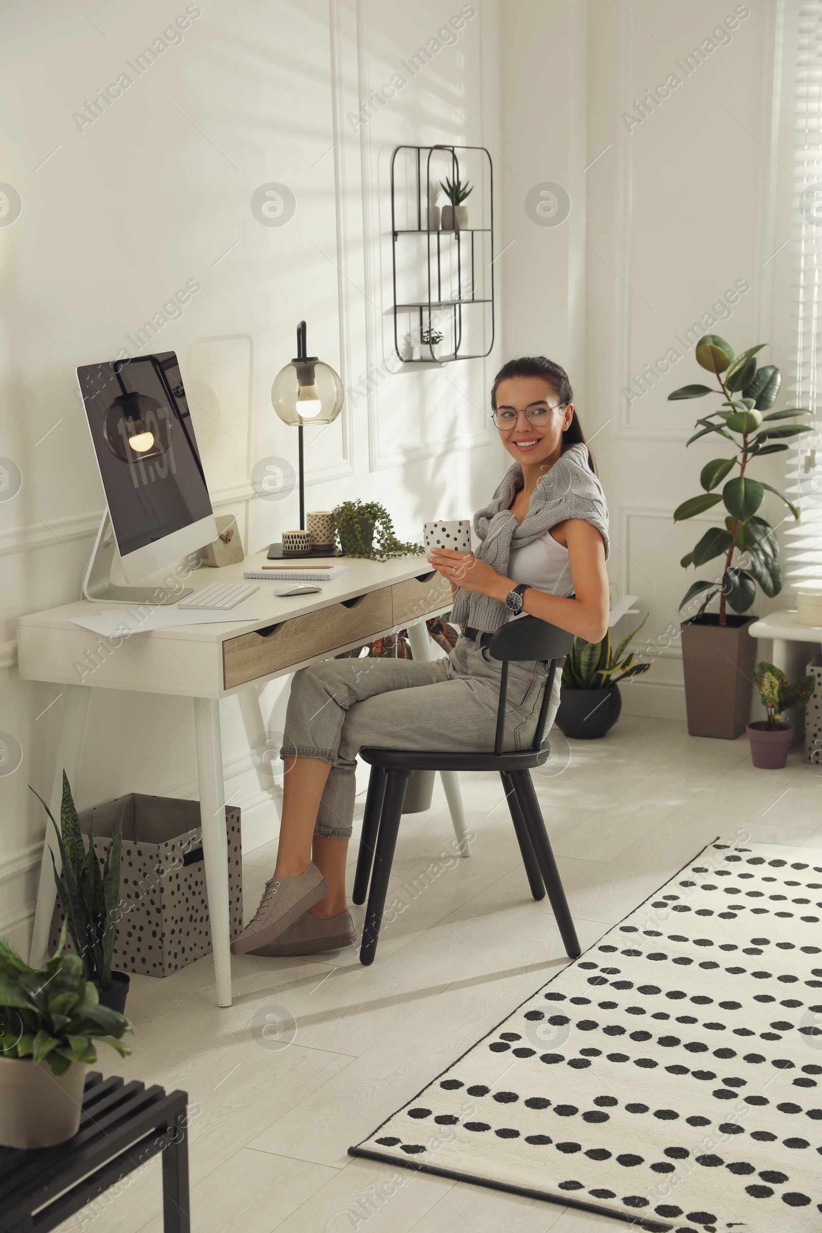 Photo of Young woman with cup of tea working at table in light room. Home office