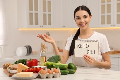 Photo of Happy woman holding notebook with words Keto Diet near different products in kitchen