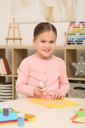 Cute little girl with glue stick and colorful paper at desk in room. Home workplace