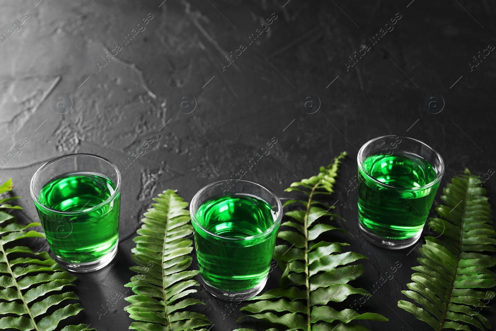 Photo of Absinthe in shot glasses and green leaves on black table, space for text. Alcoholic drink