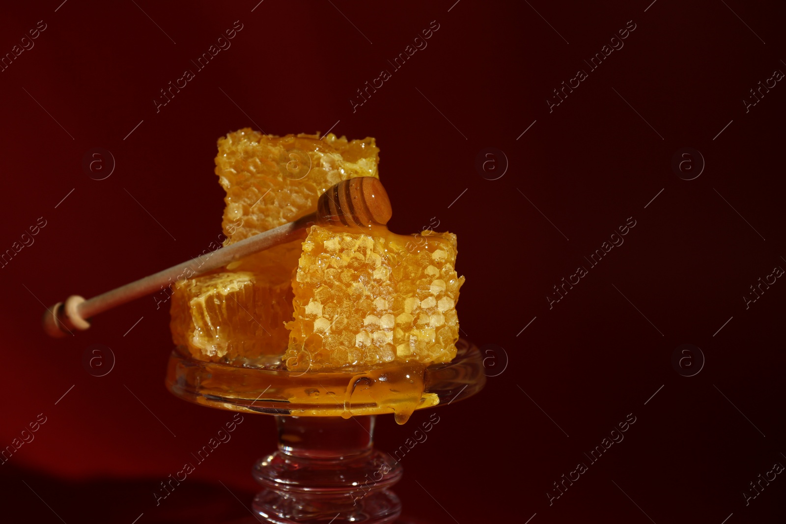 Photo of Natural honeycombs and wooden dipper on glass stand against burgundy background, space for text