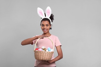 Photo of Happy African American woman in bunny ears headband holding wicker basket with Easter eggs on gray background