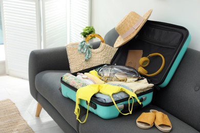 Photo of Open suitcase full of clothes, shoes and summer accessories on sofa in room