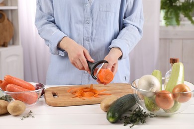 Photo of Woman peeling fresh carrot at white wooden table indoors, closeup